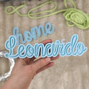 Telaio per tricotin celeste con scritta Leonardo tenuto in mano con creazioni a tricotin sullo sfondo