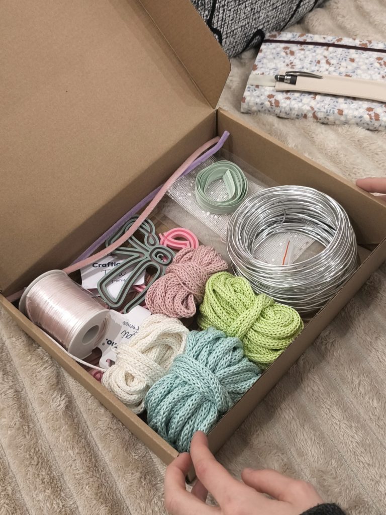 una scatola di cartone aperta con dentro vari materiali creativi: filo di alluminio, tubolare a tricotin, nastri, telai filoforma