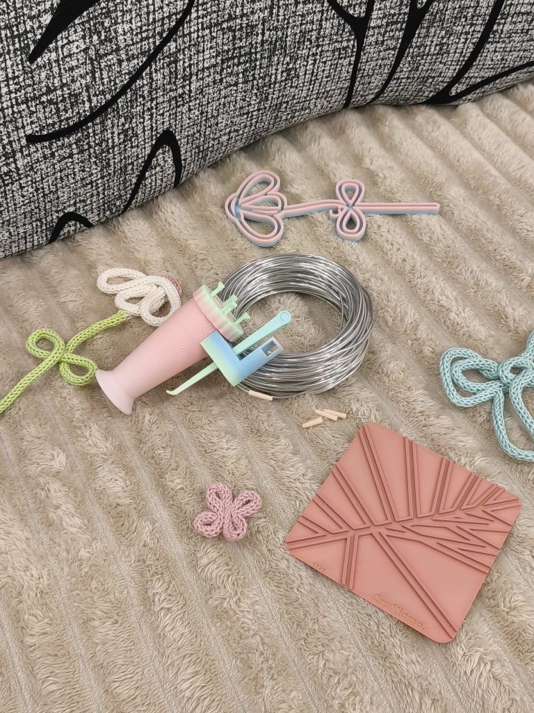 un'insieme di materiali creativi per la tecnica del tricotin: filo di alluminio, tappini per filo di alluminio, telaio base, un mulitnetto "Tina" e delle creazioni fatte a tricotin sullo sfondo